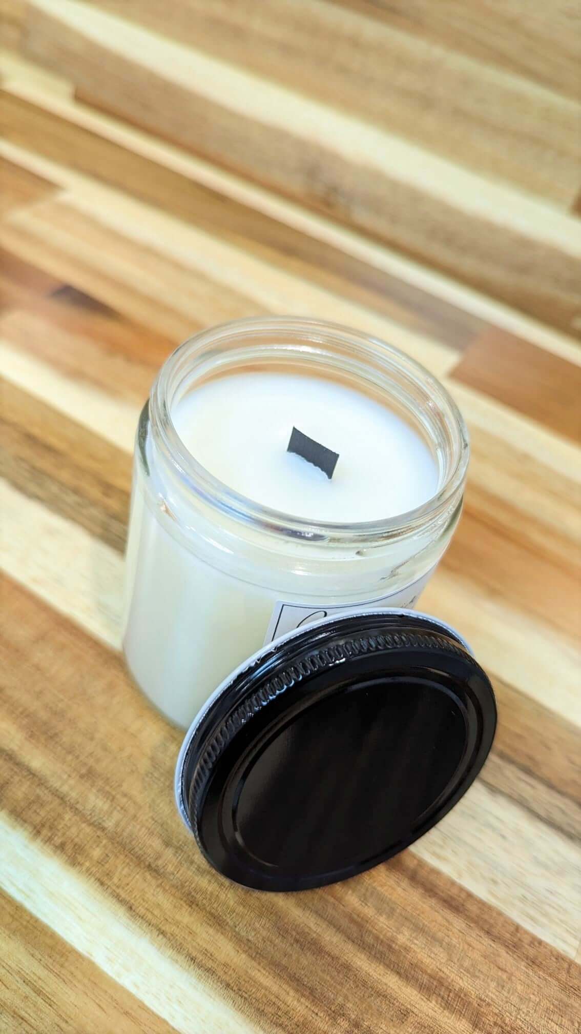 An angled picture of a candle filled jar with the black lid slanted against the side of the jar. The opened jar shows a centered black wooden wick.